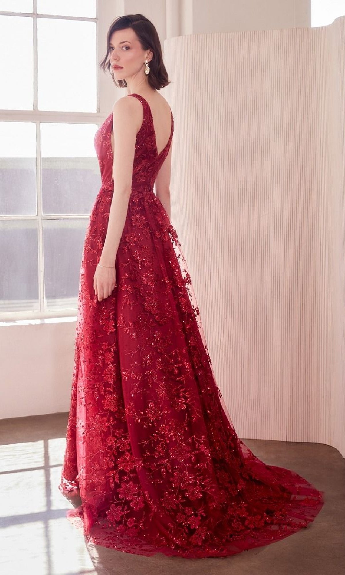 A woman stands indoors wearing the Amoris T606 Embroidered-Lace Long Prom Dress, featuring sleeveless styling, floor-length floral embroidery, and sequin details, looking over her shoulder.